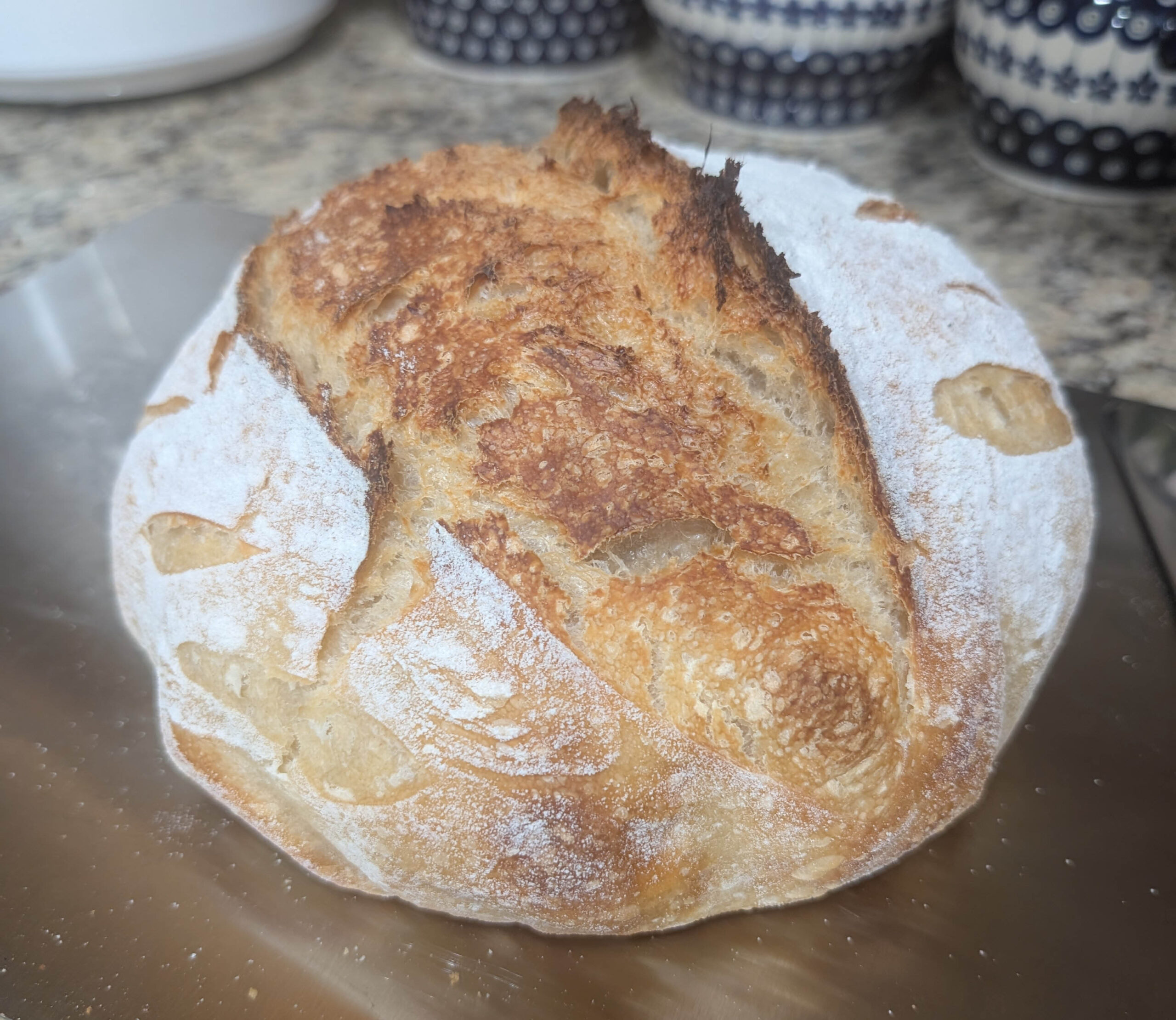 Freshly baked sourdough bread loaf on a stainless steel cutting board with polish pottery canisters in the background.